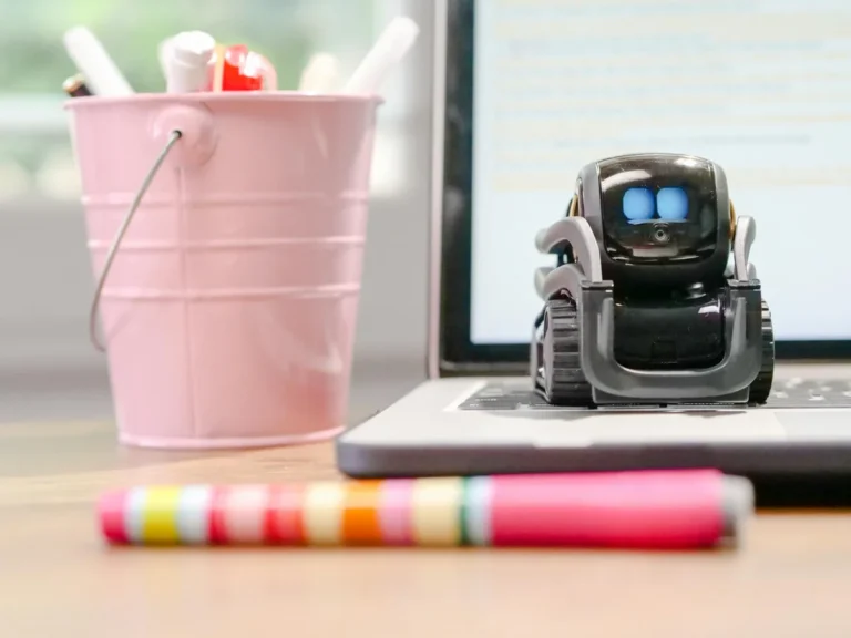 Por qué tu IA olvida lo que le dijiste: la ventana de contex... — A small robot sits on a laptop keyboard beside a pink pen holder, symbolizing te