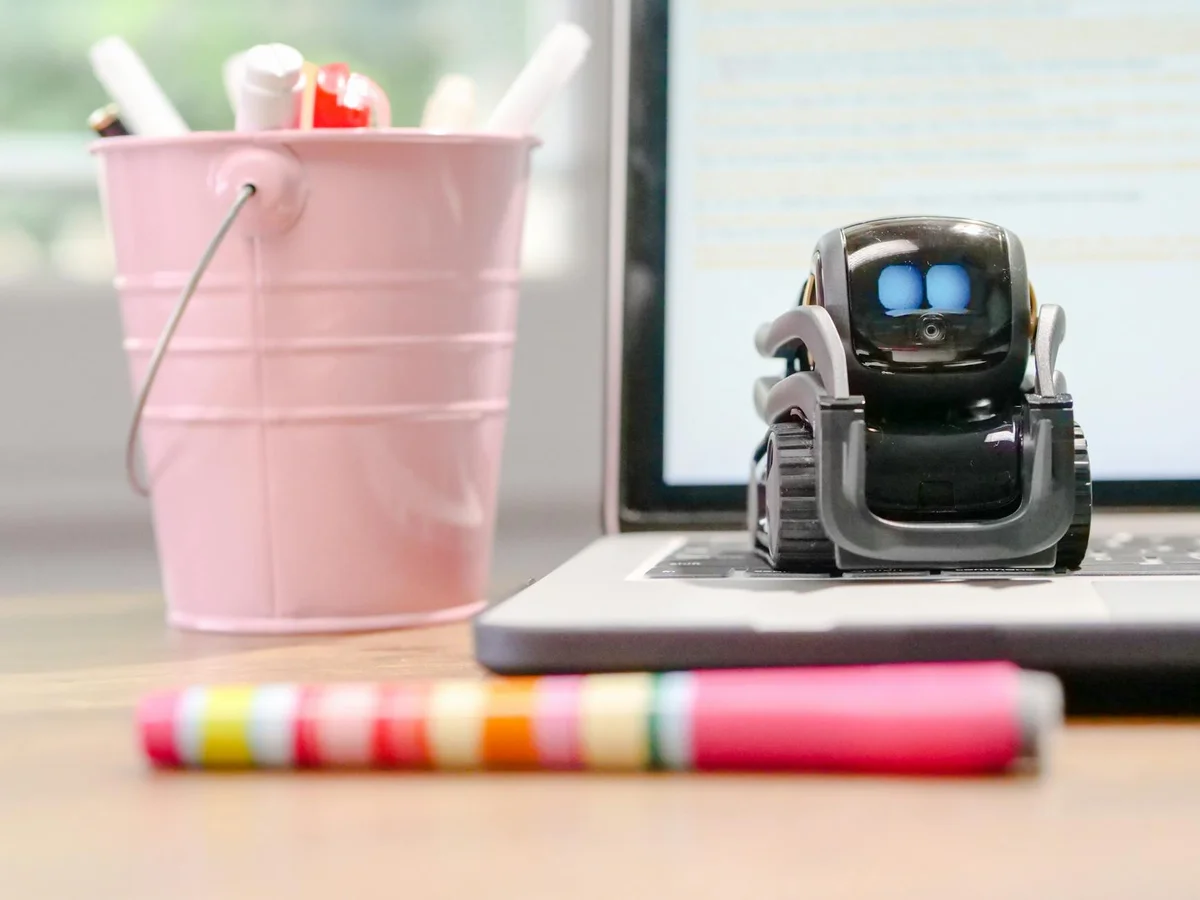 Por qué tu IA olvida lo que le dijiste: la ventana de contex... — A small robot sits on a laptop keyboard beside a pink pen holder, symbolizing te