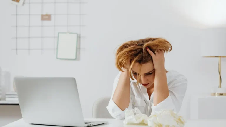 Frustrated woman in white shirt at desk with laptop, feeling overwhelmed.