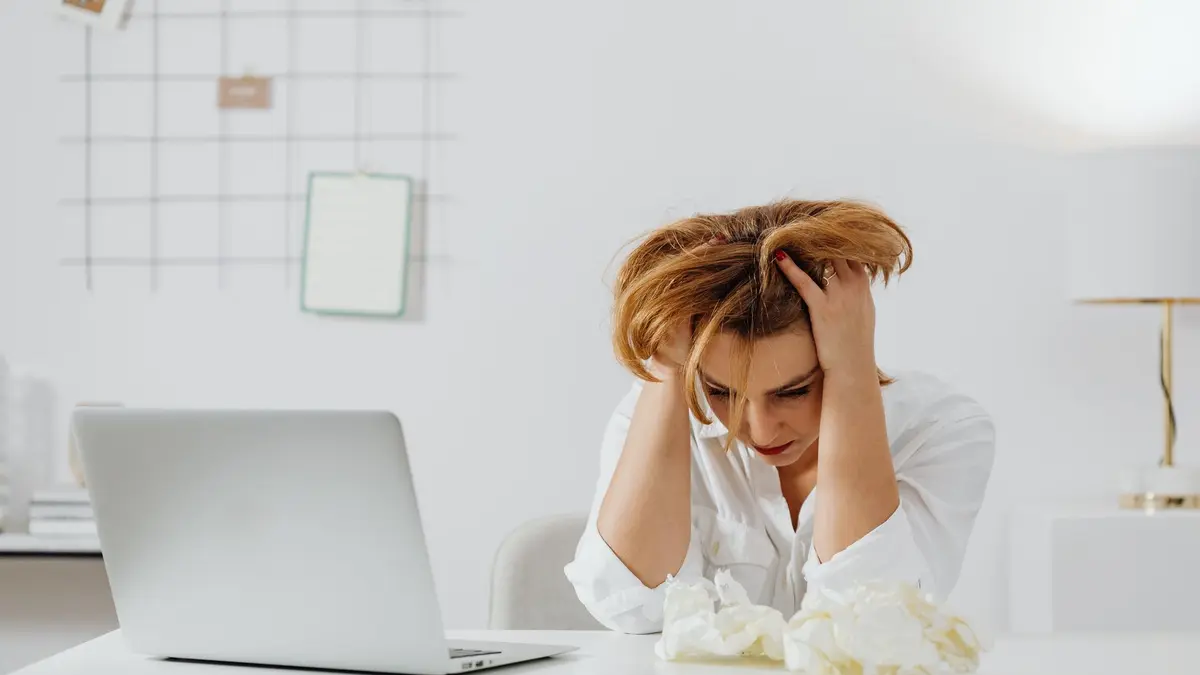 Frustrated woman in white shirt at desk with laptop, feeling overwhelmed.