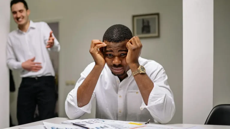 Tensed businessman at desk while coworker is laughing. Office stress concept.