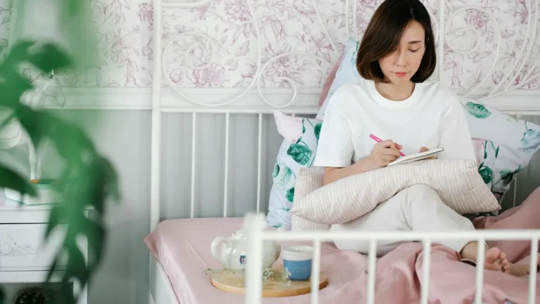 An Asian woman sitting on a bed, journaling in a serene and cozy bedroom environment.