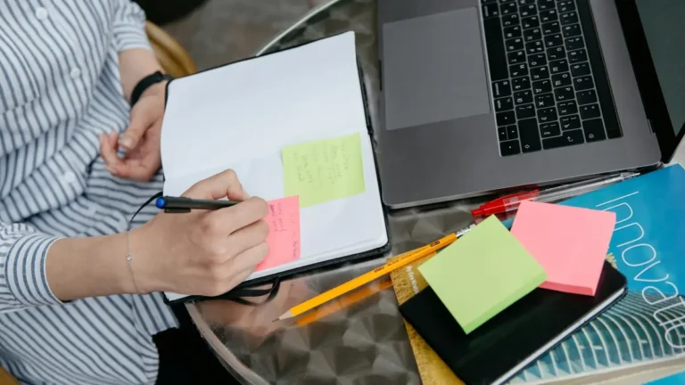 Person writing in notebook with laptop and sticky notes on a desk. Creative and organized workspace.