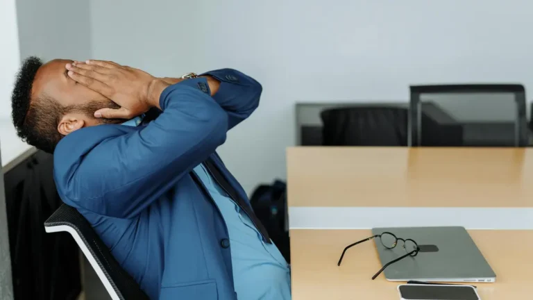 A stressed businessman in a suit covers his face while sitting at a desk with office tools.