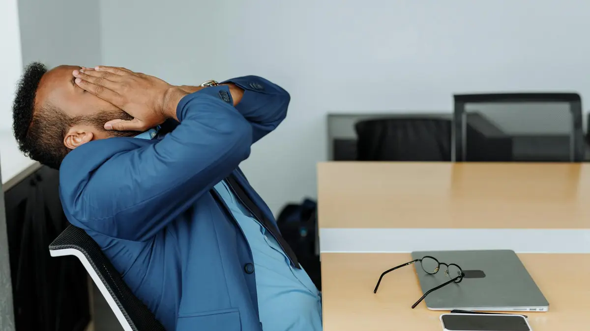 A stressed businessman in a suit covers his face while sitting at a desk with office tools.