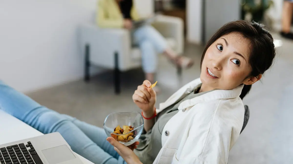 Casual scene of a woman eating chips while working on a laptop in an office setting.