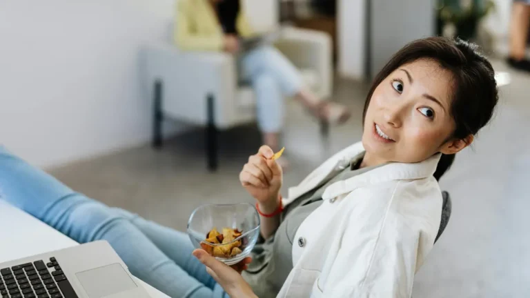 Casual scene of a woman eating chips while working on a laptop in an office setting.