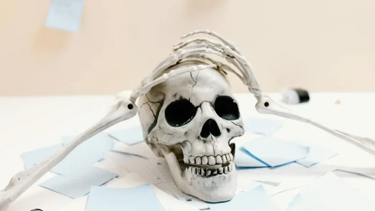 A skeleton rests on a desk with papers, symbolizing exhaustion and overwork.