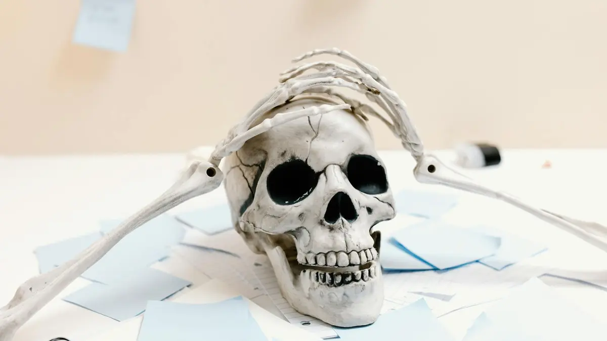 A skeleton rests on a desk with papers, symbolizing exhaustion and overwork.