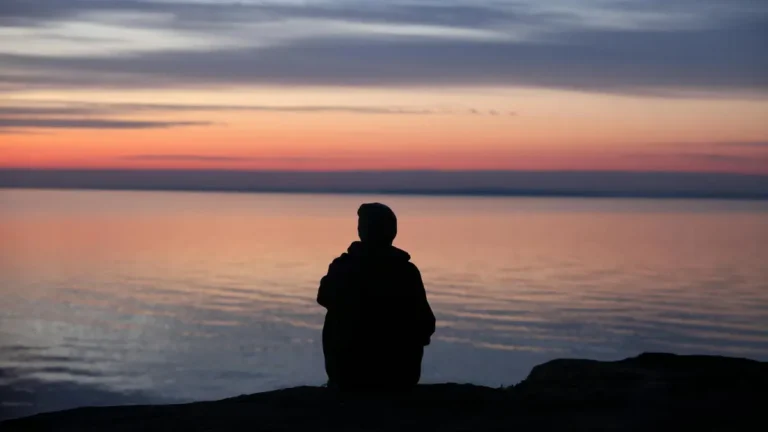 A contemplative silhouette of a person sitting by the calm sea during a beautiful sunset.