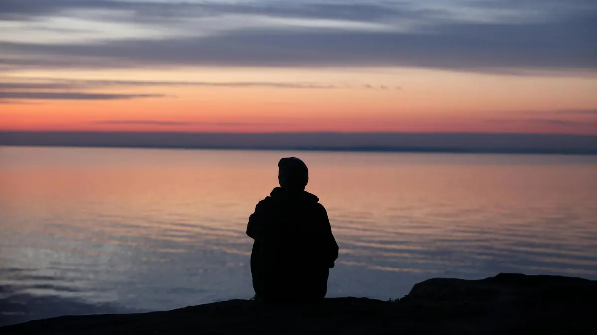 A contemplative silhouette of a person sitting by the calm sea during a beautiful sunset.