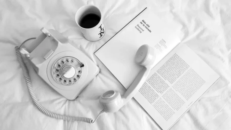 A cozy scene with a vintage rotary phone, coffee, and book on a white bed.
