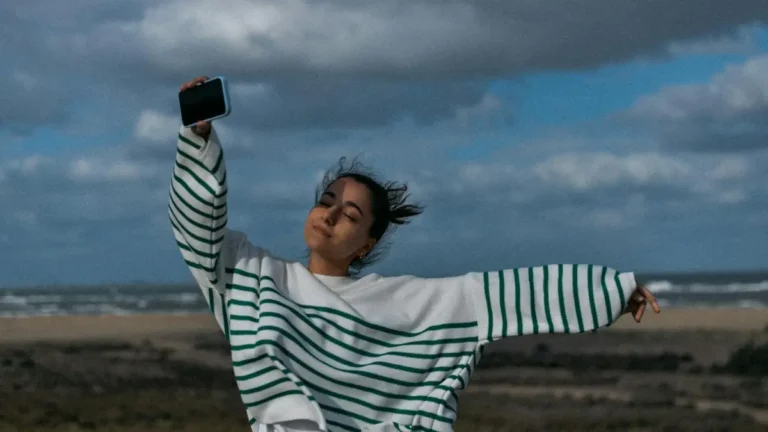Woman joyfully standing on a windy beach, holding a smartphone with eyes closed.